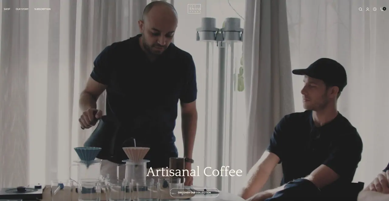 Two men in a modern cafe setting, one pouring coffee from a kettle into a dripper, the other observing, with a caption that reads "artisanal coffee.