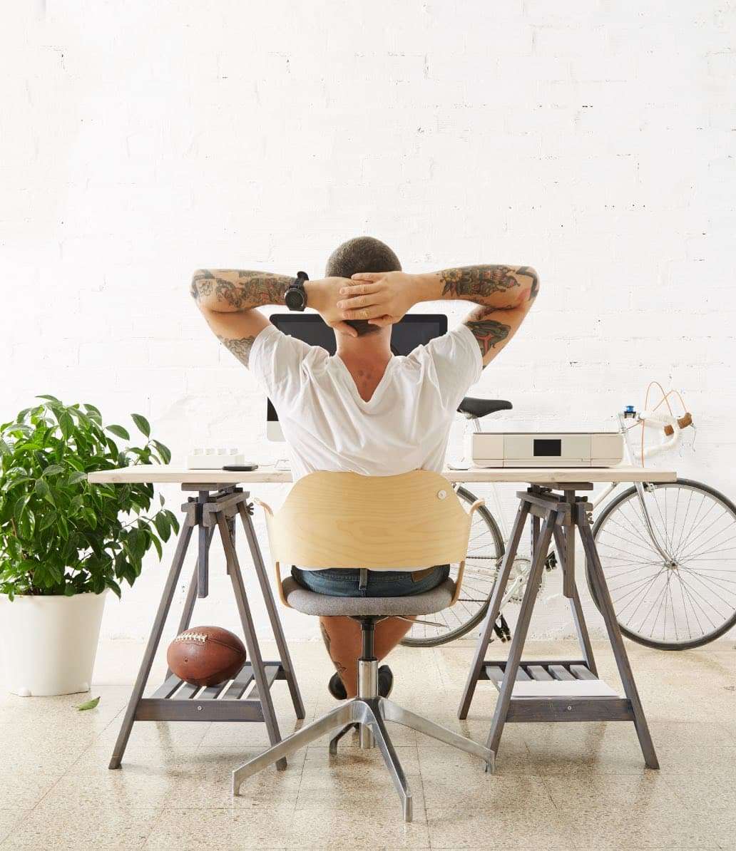 A person sits relaxed at a modern workstation with hands clasped behind head, facing a white wall. desk has computer and plant, with a bicycle leaning nearby.