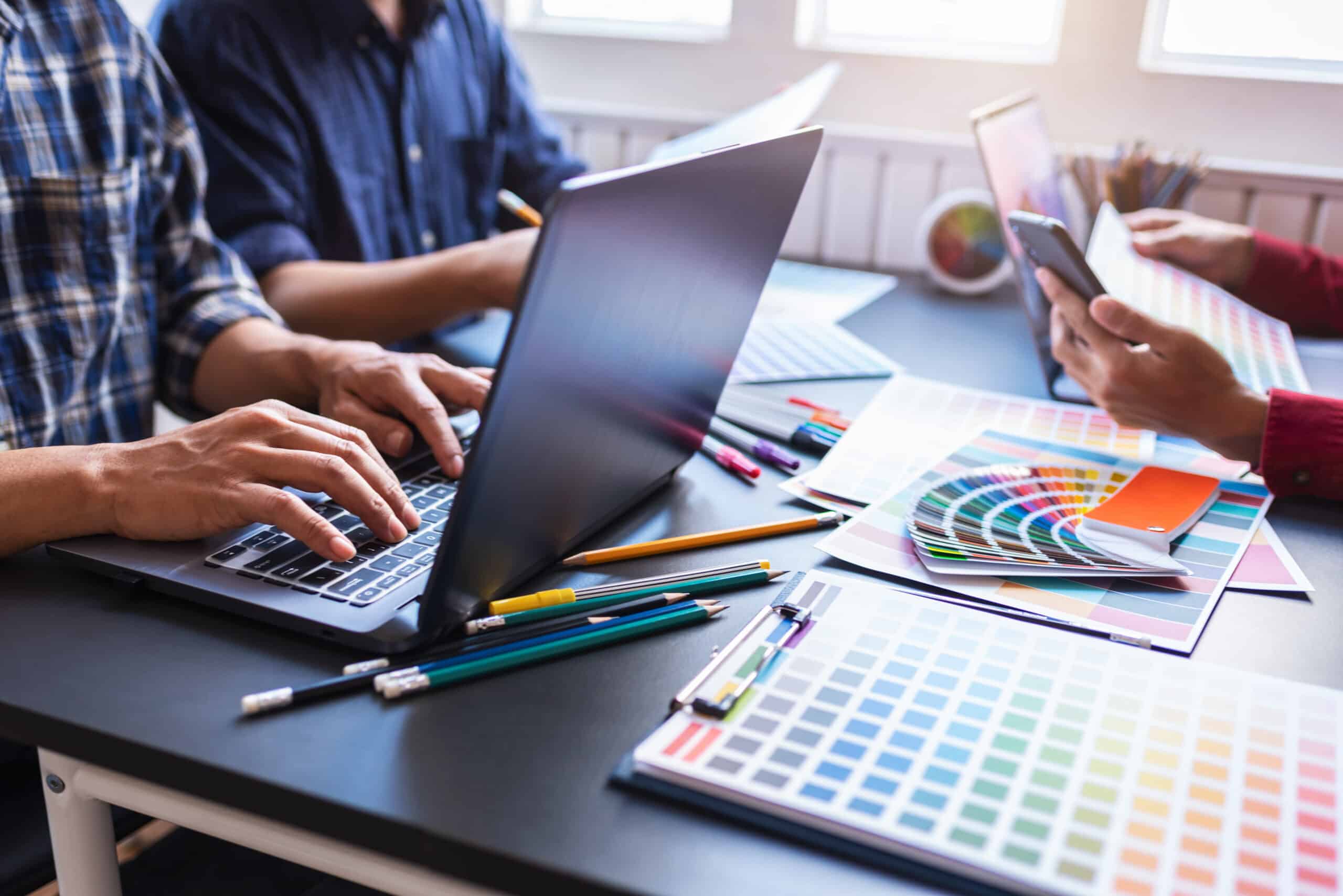 A creative team collaborates on web design at a desk filled with colour swatches, pencils, laptops, and design tools, selecting palettes and layout concepts for a digital project.