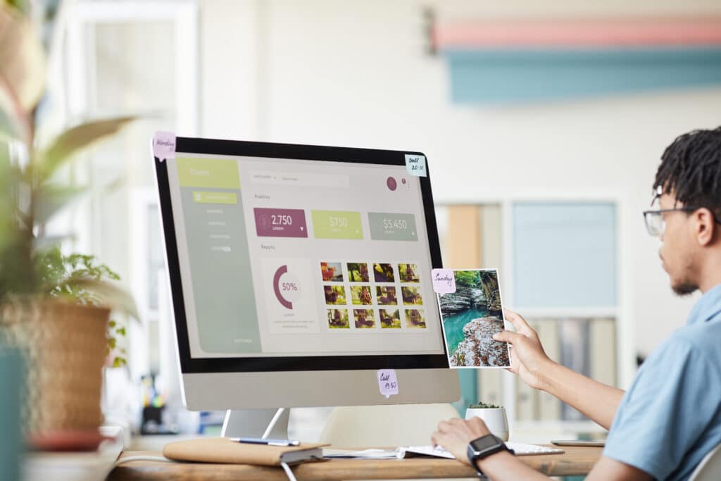 A creative professional reviews a printed photograph while working on a desktop displaying website analytics and media gallery data in a bright, modern workspace.