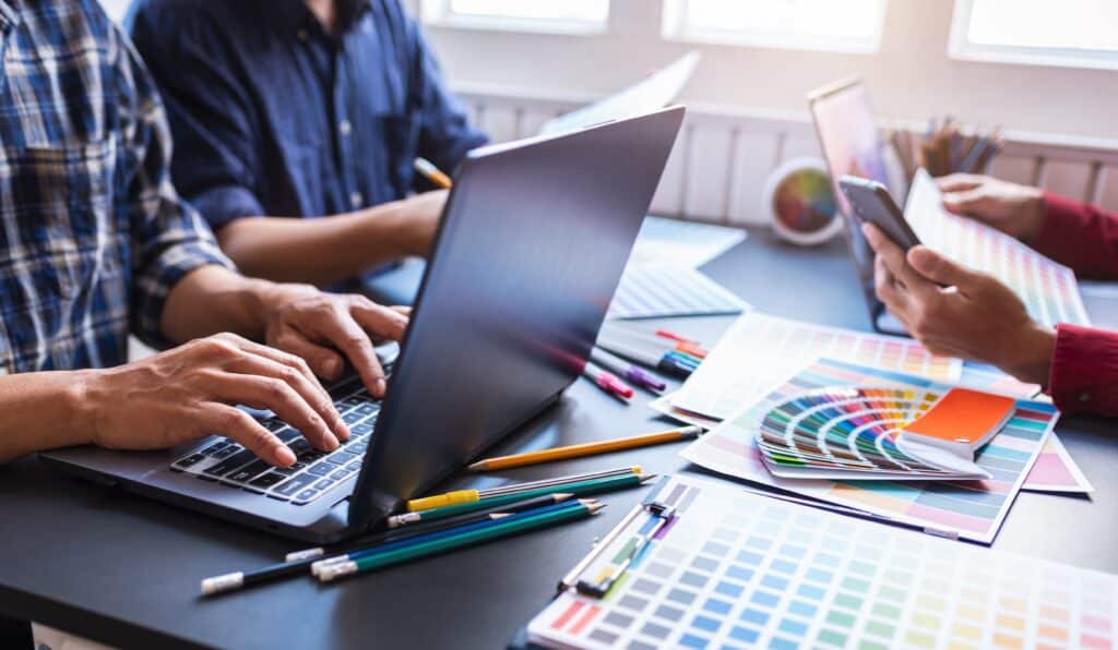 A creative team collaborating at a desk filled with laptops, colour swatches, pencils, and design tools, actively working on a professional web design project.