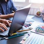 A creative team collaborating at a desk filled with laptops, colour swatches, pencils, and design tools, actively working on a professional web design project.
