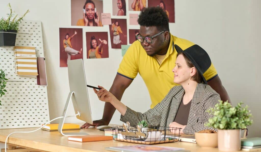 A web designer and a creative team member collaborating at a desktop in a design studio, reviewing visual content and layout elements for a digital project.