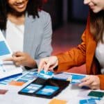 Two female designers brainstorming over a web designer layout using paper mockups and wireframes on a table, planning user interface and structure for a digital project.