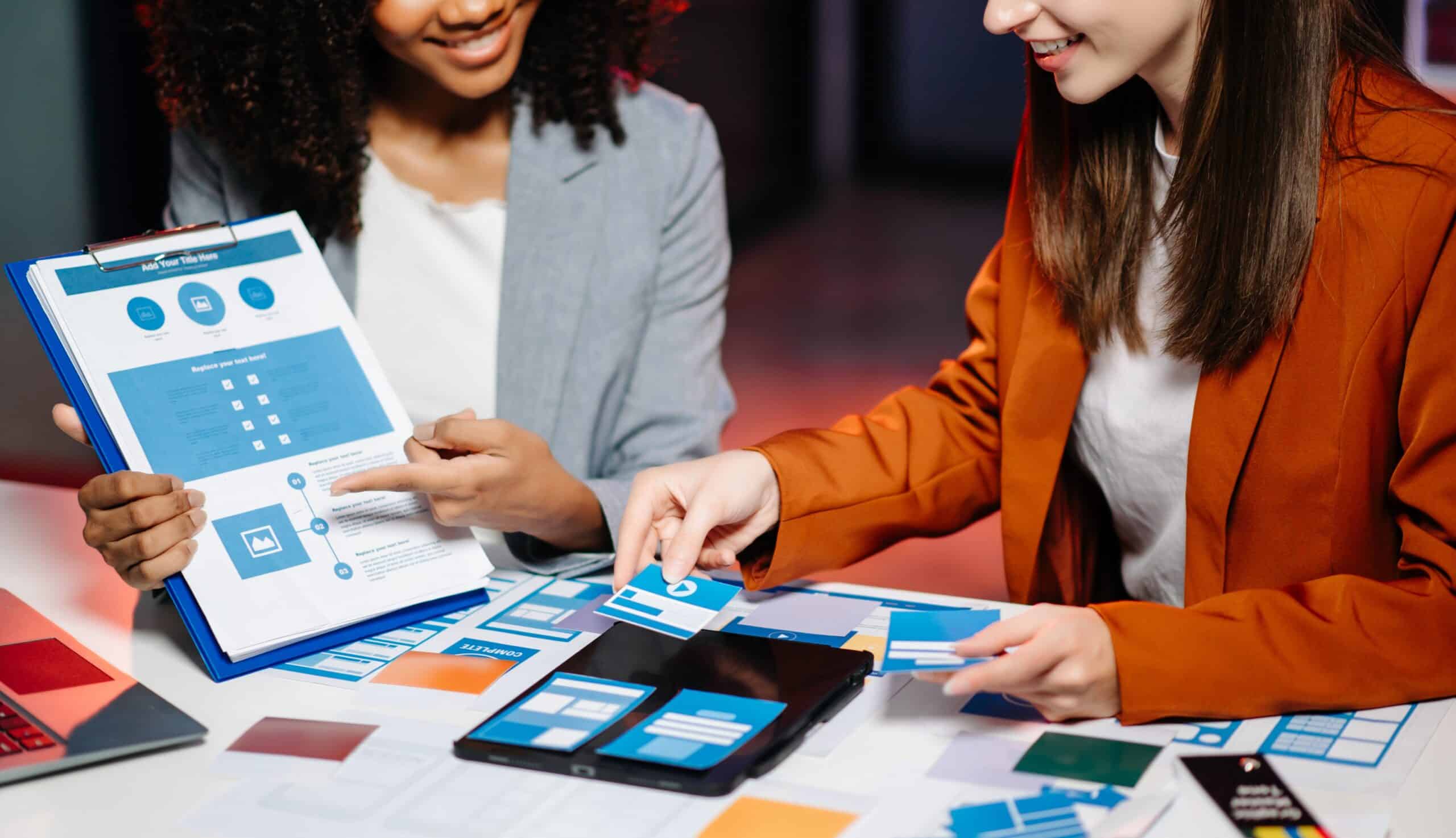 Two female designers brainstorming over a web designer layout using paper mockups and wireframes on a table, planning user interface and structure for a digital project.