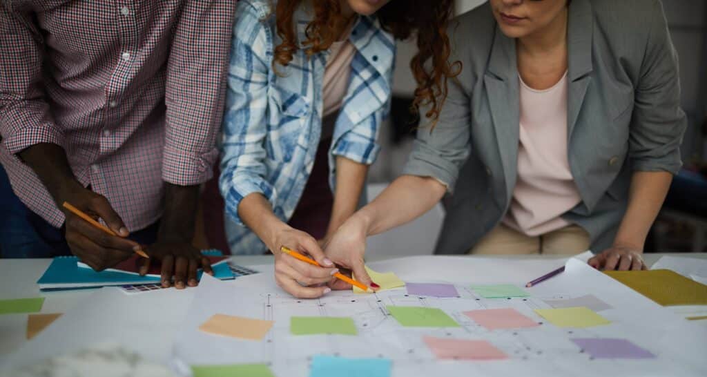 A business team discusses project management strategies at a whiteboard covered with flowcharts, graphs, sticky notes, and planning documents in a modern office setting.